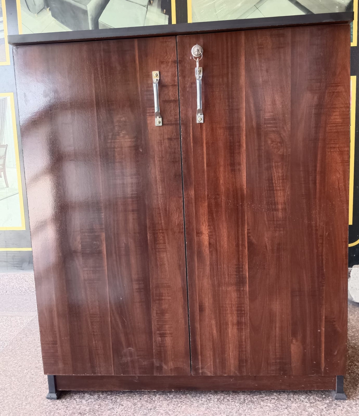 A wooden cabinet with two doors, featuring a dark reddish-brown wood grain finish, silver metal handles, and a black top surface, photographed in an indoor setting.